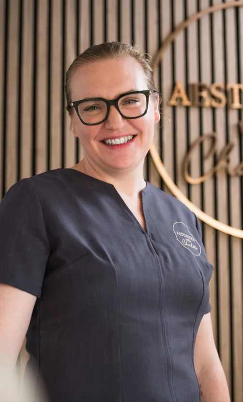Charlotte smiling in front of her gold lettered sign on the slatted wall of the clinic